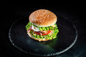 Big tasty burger with grilled meat, cheese, tomato, salad, onion rings and fried eggs on a black stone background