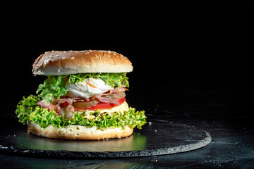 Big tasty burger with grilled meat, cheese, tomato, salad, onion rings and fried eggs on a black stone background