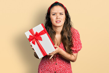 Young hispanic girl holding gift clueless and confused expression. doubt concept.