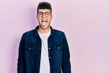 Young hispanic man wearing casual clothes and glasses sticking tongue out happy with funny expression. emotion concept.