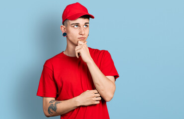 Young caucasian boy with ears dilation wearing delivery uniform and cap serious face thinking about question with hand on chin, thoughtful about confusing idea