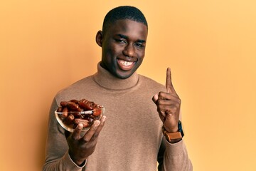 Young african american man holding bowl with dates smiling with an idea or question pointing finger with happy face, number one