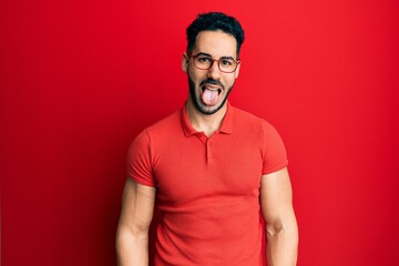 Young hispanic man wearing casual clothes and glasses sticking tongue out happy with funny expression. emotion concept.