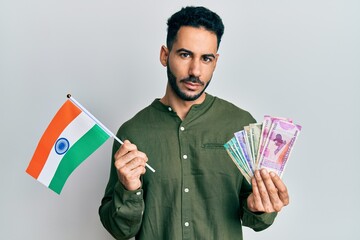 Young hispanic man holding india flag and rupee banknotes relaxed with serious expression on face. simple and natural looking at the camera.