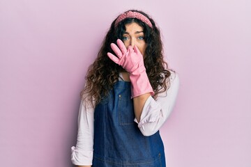 Young brunette woman with curly hair wearing cleaner apron and gloves smelling something stinky and disgusting, intolerable smell, holding breath with fingers on nose. bad smell