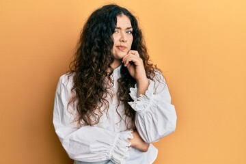 Young brunette woman with curly hair wearing casual clothes thinking concentrated about doubt with finger on chin and looking up wondering