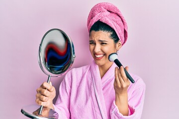 Young hispanic woman wearing shower bathrobe using makeup brush clueless and confused expression....