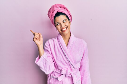 Young hispanic woman wearing shower towel cap and bathrobe with a big smile on face, pointing with hand and finger to the side looking at the camera.