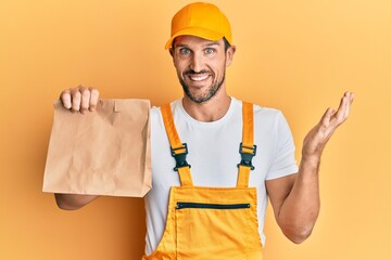 Young handsome delivery man holding take away paper bag celebrating achievement with happy smile and winner expression with raised hand