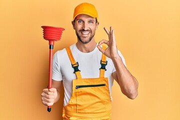 Young handsome man wearing plumber uniform holding toilet plunger doing ok sign with fingers,...