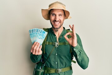 Handsome man with beard wearing explorer hat holding 100 brazilian reals doing ok sign with fingers, smiling friendly gesturing excellent symbol