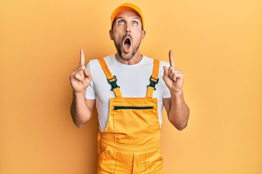 Young handsome man wearing handyman uniform over yellow background amazed and surprised looking up and pointing with fingers and raised arms.
