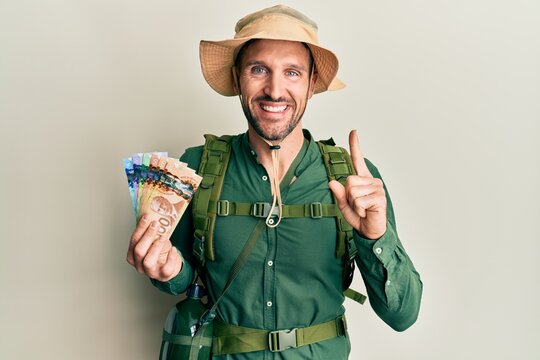Handsome man with beard wearing explorer hat holding canadian dollars smiling with an idea or question pointing finger with happy face, number one