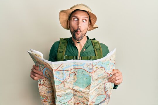 Handsome man with beard wearing explorer hat holding map making fish face with mouth and squinting eyes, crazy and comical.