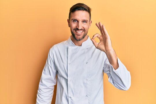 Handsome man with beard wearing professional cook uniform smiling positive doing ok sign with hand and fingers. successful expression. - Powered by Adobe