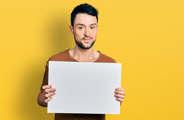 Hispanic man with beard holding blank empty banner relaxed with serious expression on face. simple and natural looking at the camera.