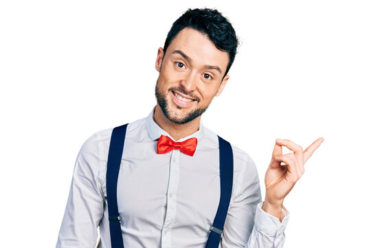 Hispanic man with beard wearing hipster look with bow tie and suspenders with a big smile on face, pointing with hand and finger to the side looking at the camera.