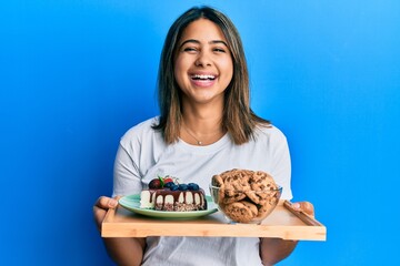 Young latin woman holding cake sweets and cookies smiling and laughing hard out loud because funny crazy joke.