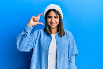 Young latin woman wearing cute wool cap smiling and confident gesturing with hand doing small size sign with fingers looking and the camera. measure concept.