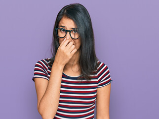 Beautiful asian young woman wearing casual clothes and glasses smelling something stinky and disgusting, intolerable smell, holding breath with fingers on nose. bad smell