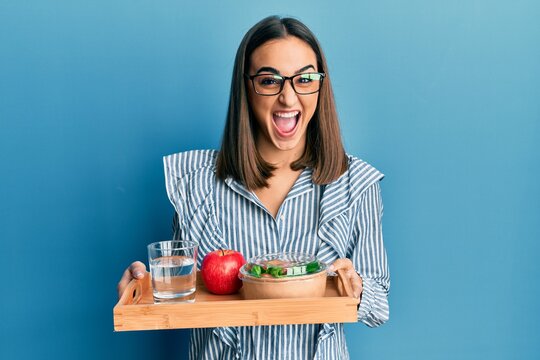 Young brunette girl holding tray with healthy lunch celebrating crazy and amazed for success with open eyes screaming excited.