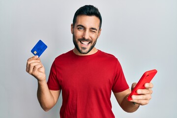 Young hispanic man holding smartphone and credit card winking looking at the camera with sexy...