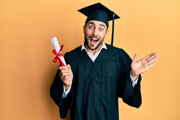 Young hispanic man wearing graduation robe holding diploma celebrating achievement with happy smile and winner expression with raised hand - obrazy, fototapety, plakaty