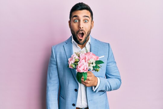 Young hispanic businessman wearing business jacket holding flowers scared and amazed with open mouth for surprise, disbelief face