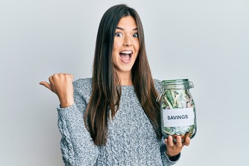 Young brunette woman holding jar with savings pointing thumb up to the side smiling happy with open...