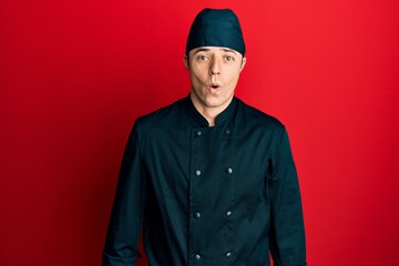 Handsome young man wearing professional cook uniform and hat afraid and shocked with surprise expression, fear and excited face.