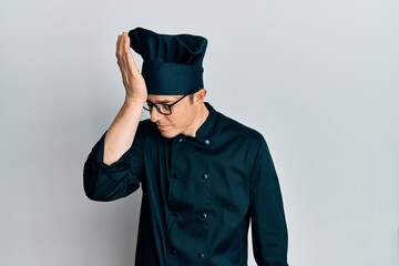 Handsome young man wearing professional cook uniform and hat surprised with hand on head for mistake, remember error. forgot, bad memory concept.