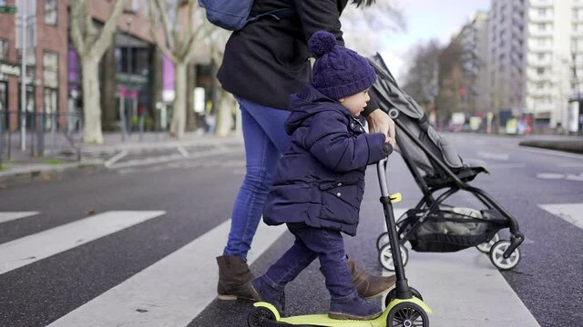 Child and mother crossing street on crosswalk. Kid riding scooter with parent