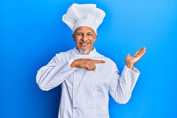 Middle age grey-haired man wearing professional cook uniform and hat amazed and smiling to the camera while presenting with hand and pointing with finger.