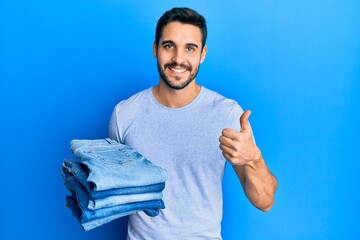 Young hispanic man holding stack of folded jeans smiling happy and positive, thumb up doing...