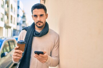 Young hispanic man with serious expression using smartphone and drinking take away coffee at the city.