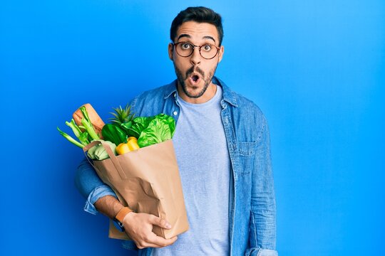 Young hispanic man holding paper bag with bread and groceries scared and amazed with open mouth for surprise, disbelief face