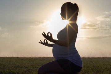 A healthy pregnant woman doing yoga and meditation in nature with a sunset silhouette. Prenatal health and wellness.