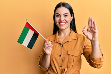 Young brunette woman holding united arab emirates flag doing ok sign with fingers, smiling friendly gesturing excellent symbol