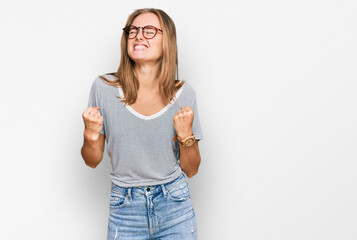 Fototapeta na wymiar Beautiful young blonde woman wearing casual clothes and glasses very happy and excited doing winner gesture with arms raised, smiling and screaming for success. celebration concept.