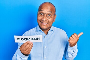 Middle age latin man holding paper with blockchain message pointing thumb up to the side smiling happy with open mouth