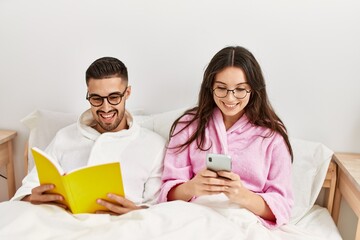 Young hispanic couple reading book and using smartphone at bed.