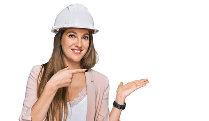 Young blonde woman wearing architect hardhat amazed and smiling to the camera while presenting with hand and pointing with finger.