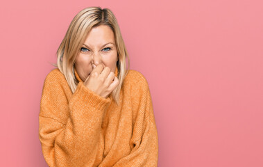 Middle age caucasian woman wearing casual winter sweater smelling something stinky and disgusting, intolerable smell, holding breath with fingers on nose. bad smell