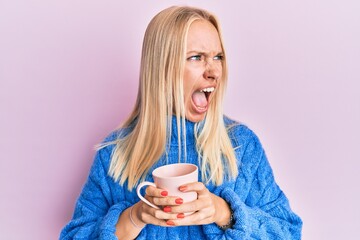Young blonde girl wearing winter sweater and drinking a cup of hot coffee angry and mad screaming...
