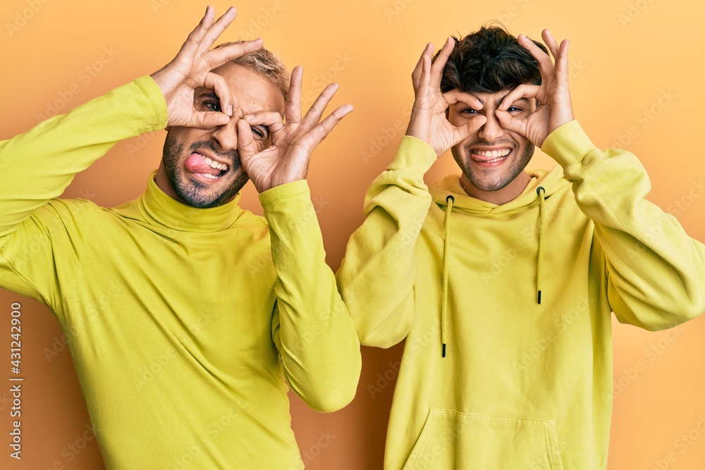 Wall mural Homosexual gay couple standing together wearing yellow clothes doing ok gesture like binoculars sticking tongue out, eyes looking through fingers. crazy expression.