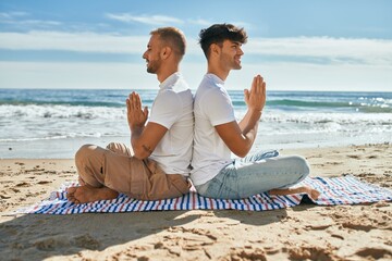 Young gay couple doing yoga sitting at the beach.