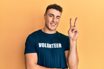 Hispanic young man wearing volunteer t shirt smiling with happy face winking at the camera doing...