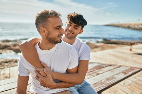 Young gay couple smiling happy sitting on the bench at the beach promenade.