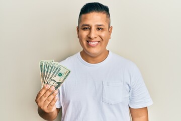 Young latin man holding 20 dollars banknotes looking positive and happy standing and smiling with a...