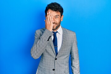 Handsome man with beard wearing business suit and tie covering one eye with hand, confident smile on face and surprise emotion.